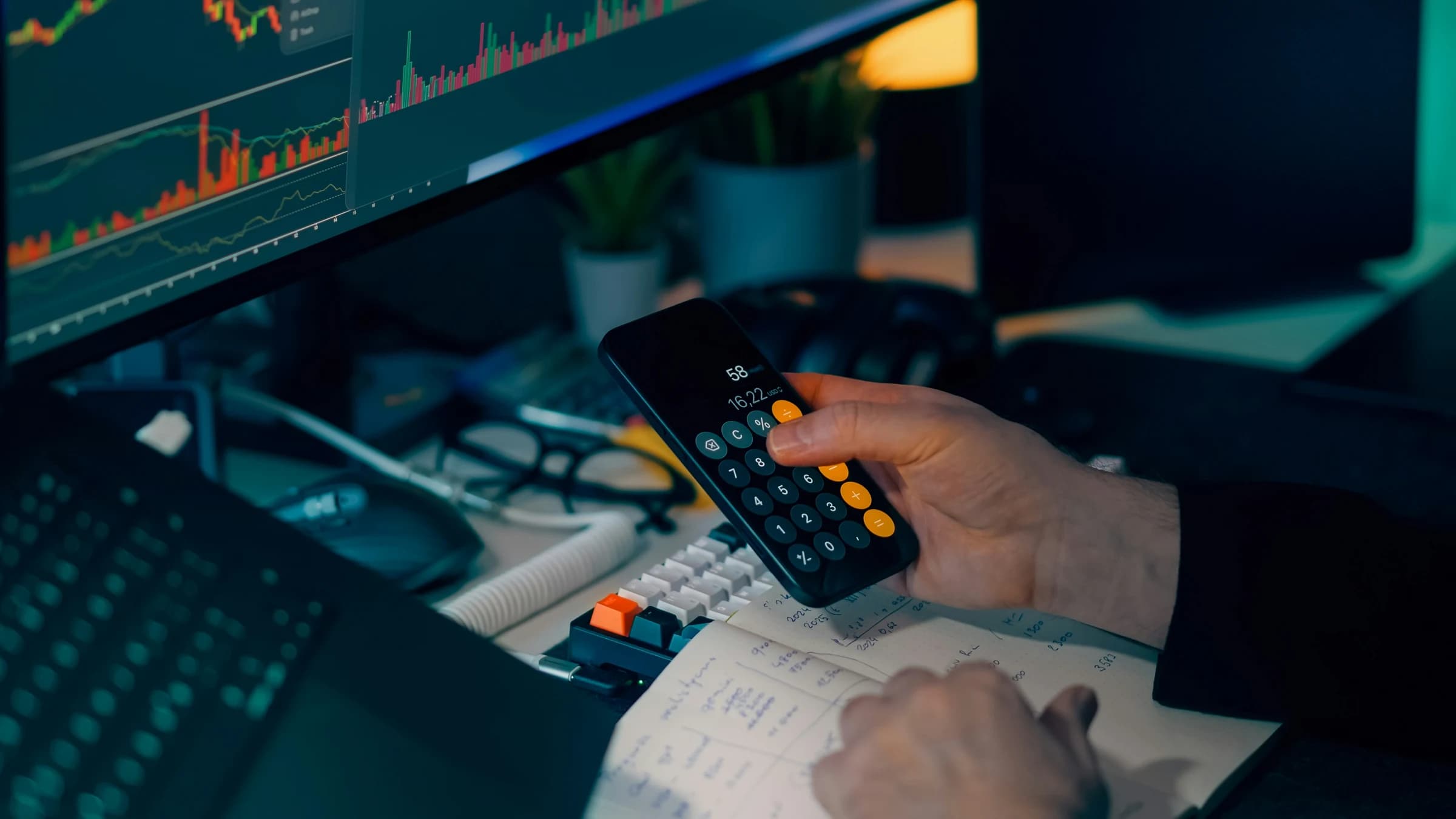 Person calculating investment returns using a calculator with stock market charts on screen